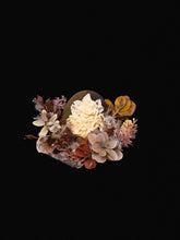 The Magnolia Birth Flower Ring by CAMELLIA, featuring an intricate ivory rose reminiscent of handcrafted jewelry, is artfully set amidst dried brown and orange leaves against a black background.